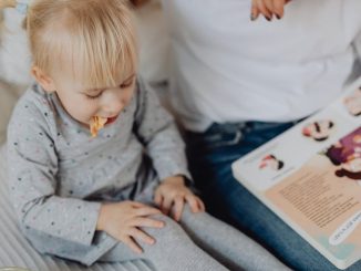 Cute toddler with ponytails enjoying a snack next to a book and parent. Cozy indoor scene.