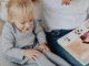 Cute toddler with ponytails enjoying a snack next to a book and parent. Cozy indoor scene.